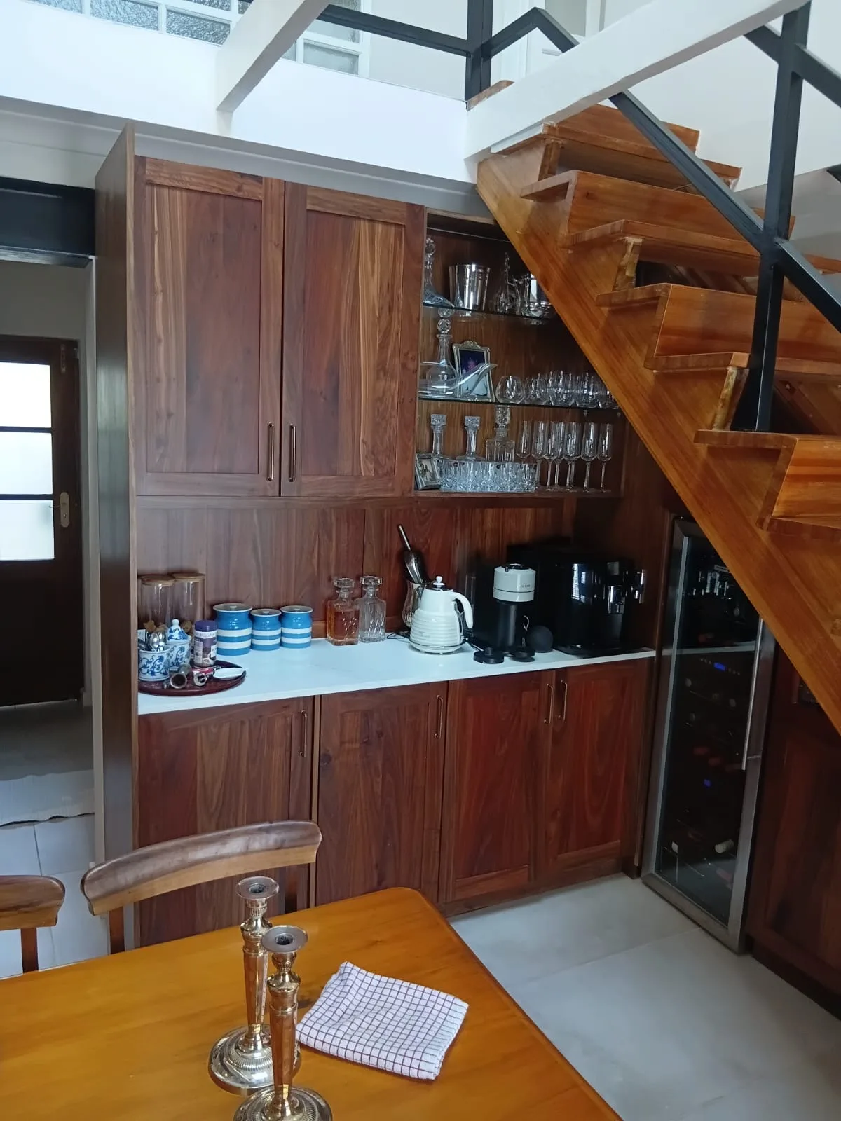 Under-Stair Bar in Solid Timber — The dead space under an open timber staircase turned into a bar: glass shelving for glassware, a coffee station on a white top, and a wine fridge built into the run, all in a rich figured timber that matches the treads.