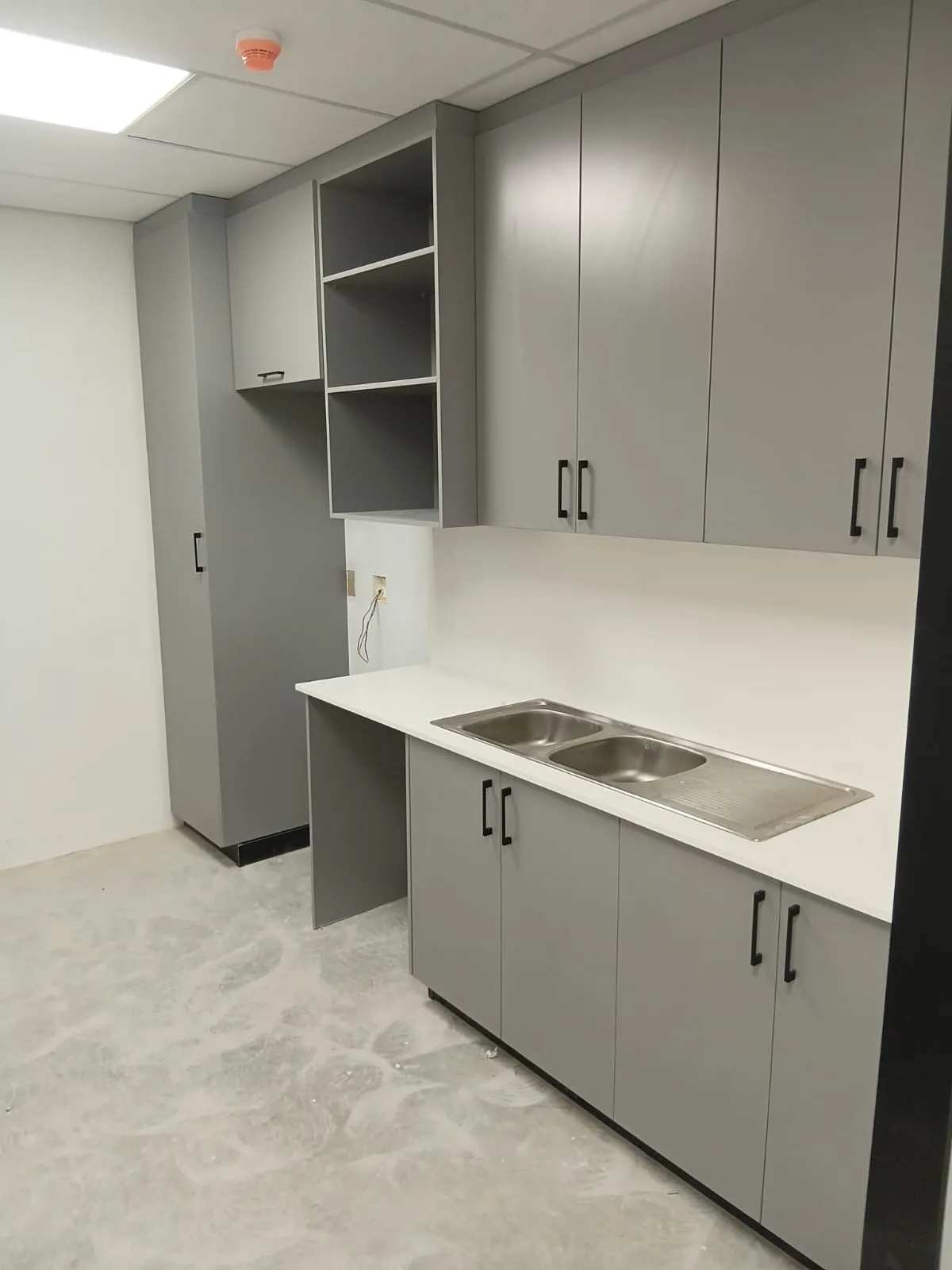 Grey Matt Office Kitchenette — A staff kitchenette fitted into an office, in matt grey with black bar handles. A double stainless sink sits in a white top, with an open shelving niche above the counter and a full-height store cupboard at the end of the run.