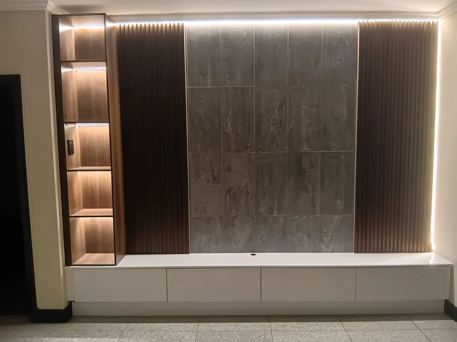Lit Media Wall with Fluted Panels — Fluted timber panels either side of a concrete-look tiled centre, with LED strip washing down the wall from a recess above. A lit display tower runs full height on the left, and the white drawer unit below floats clear of the floor.
