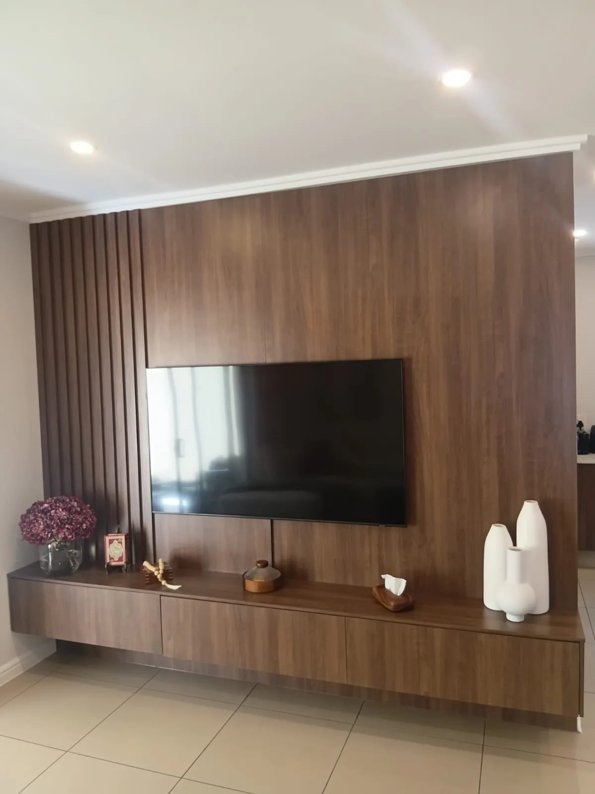 Walnut Media Wall, Floor to Ceiling — A full-height walnut panel wrapping the whole wall, with a fluted section down one side to break up the grain. The floating unit beneath it has three deep drawers and hides every cable and decoder.