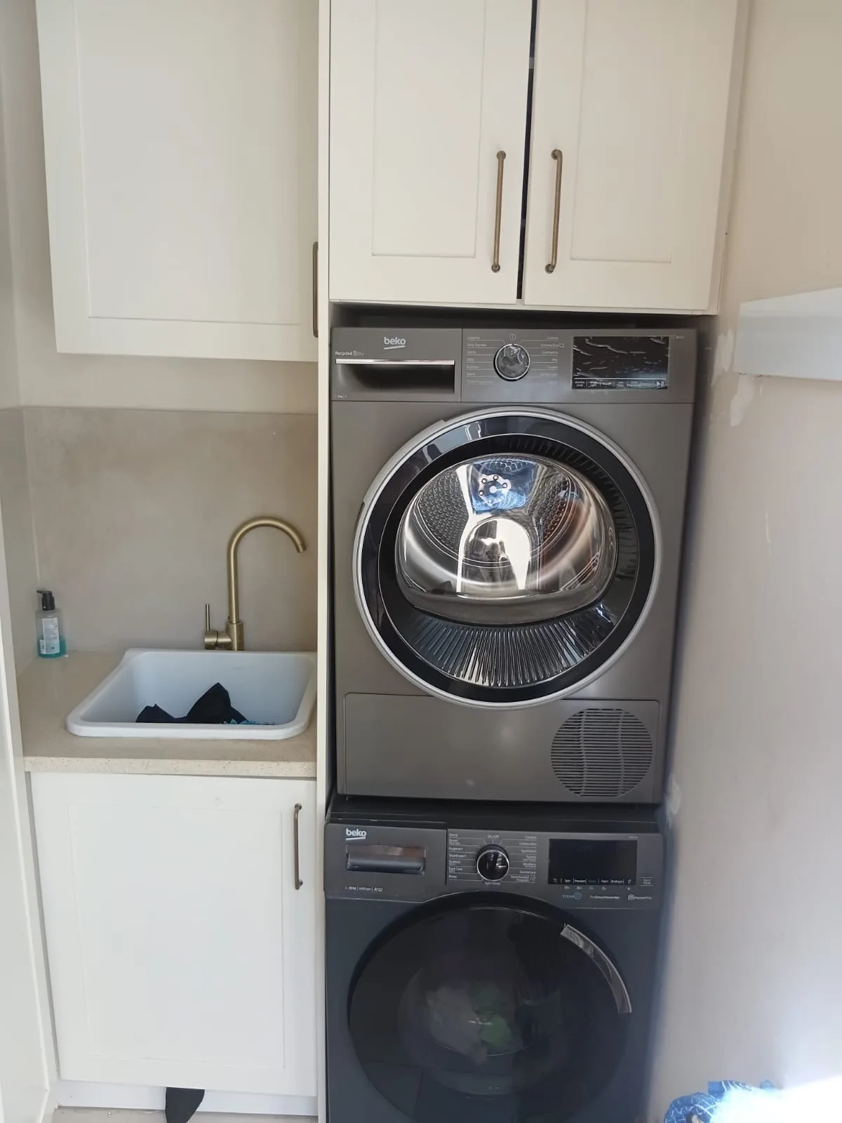 Shaker Laundry with Stacked Machines — A laundry fitted in shaker doors, with the washer and tumble dryer stacked into a single tall housing so the room keeps its floor space. Small sink with a brass tap, and cupboards above and below.