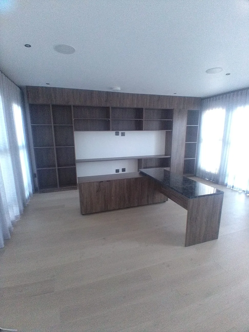 Built-In Home Office — A wall of built-in shelving and closed storage in a smoked oak finish, with an L-shaped desk that wraps into the room. The return is topped in granite and sized for a second monitor.